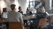 Photo of five employees looking at each other. They are gathered around a table in a meeting room